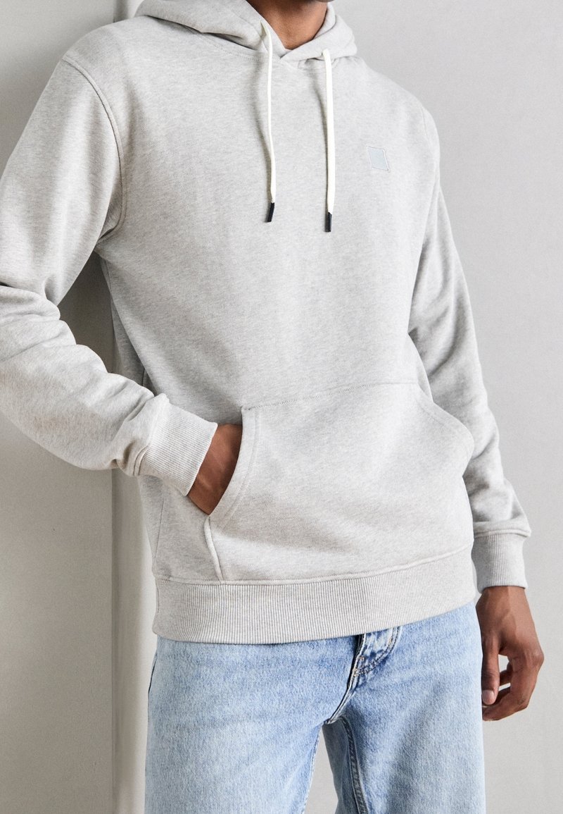 Sudadera gris claro con bolsillo canguro, capucha con cordón, puños acanalados y un pequeño logo frontal. Combinada con jeans claros azul.
