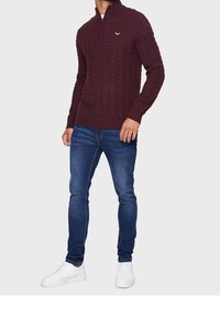 Bordeauxfarbener Strickpullover mit einem Viertelreißverschluss, der ein strukturiertes Zopfmuster und ein kleines Logo auf der Brust aufweist, kombiniert mit blauen Jeans und weißen Sneakern.
