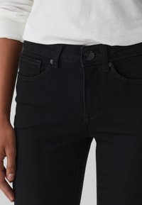 Persona vestida con jeans negros de cierre de botón y cremallera, combinados con una blusa blanca de manga larga, con la mano descansando a un lado.