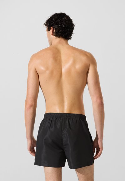 Shorts de bain noirs avec une ceinture élastique, une texture lisse et une coupe ample, montrés de dos sur un torse nu contre un fond neutre.