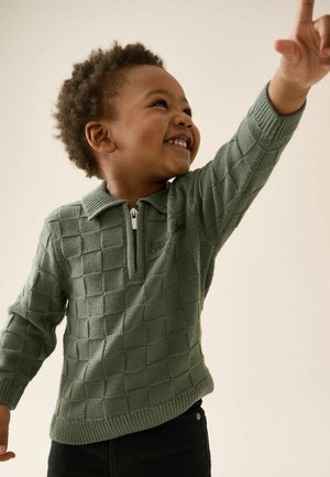 Lächelndes Kind mit grünem, strukturiertem Pullover zeigt mit der rechten Hand nach oben vor einem einfarbigen hellen Hintergrund.
