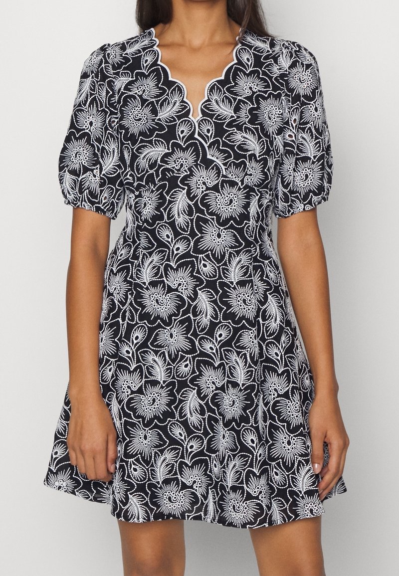 Femme portant une robe noire avec broderie florale blanche, manches bouffantes et encolure en V festonnée, montrée du cou à mi-cuisse.