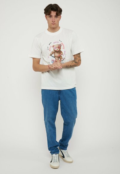 Giovane uomo con tatuaggi che indossa una maglietta bianca con grafica di un orsacchiotto, jeans blu e sneakers bianche, in piedi con le mani giunte.
