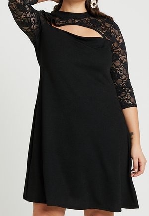 Frau trägt ein schwarzes Kleid mit Spitzen-3/4-Ärmeln und einem diagonalen Ausschnitt über der Brust, ergänzt durch große Ohrringe.