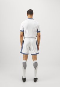 Witte voetbaloutfit met blauwe accenten, bestaande uit een shirt met korte mouwen en een korte broek. Gestreepte kniekousen worden gecombineerd met zwarte voetbalschoenen.