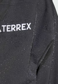 Musta vedenpitävä takki, jonka pinnalla on vesipisaroita. Suuri valkoinen "Terrex"-logo. Tasainen pinta ja tyylikäs muotoilu.