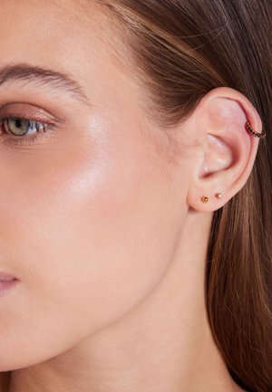 Goldene und mehrfarbige Ohrringe schmücken das Ohr eines Modells. Das Design umfasst einen Ring und zwei Steckerohrringe, die einen modernen Look betonen.