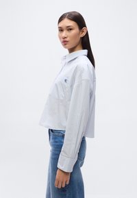 Κοντοκομμένο πουκάμισο με κουμπιά σε ανοιχτό μπλε με κάθετες ρίγες, με ένα μικρό κεντημένο λογότυπο και φαρδιά μανίκια, σε συνδυασμό με στενά τζιν.