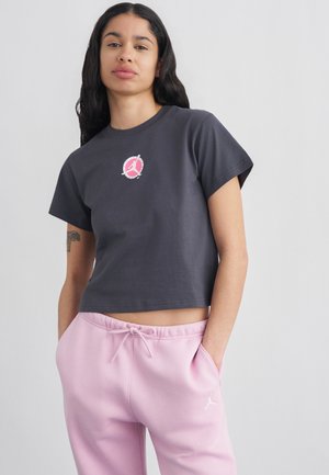 Tânără cu păr lung și închis la culoare, purtând un tricou scurt închis și pantaloni de trening roz, cu mâinile în buzunare, pe un fundal deschis.