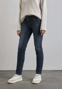 Person som bär mörkblå slim fit-jeans, en beige tröja och vita sneakers, stående på ett grått golv mot en enfärgad vägg.
