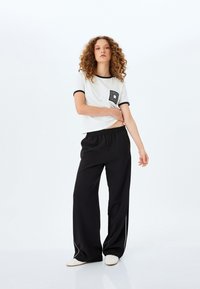 Camiseta blanca de manga corta con ribete negro y un bolsillo con patrón de diamante, combinada con pantalones negros holgados con ribete blanco.
