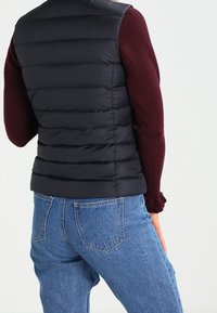 Svart pufferväst med horisontellt kviltat mönster, med hög krage och åtsittande design. Buren över en mörkröd långärmad tröja och blå jeans.