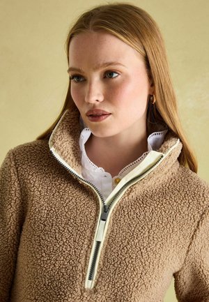 Brauner, flauschiger Fleece-Pullover mit einem Reißverschlusskragen und cremefarbenen Akzenten, über einem weißen Hemd mit Kragen und Spitzenrand getragen.