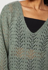 Pull en tricot vert clair avec col en V et motif ajouré, présentant une coupe décontractée et un fil texturé et doux.