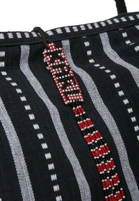 Čierna látková taška so bielymi pruhmi a korálikovým dekorom s geometrickým vzorom v červenej, čiernej a bielej farbe. Súčasťou je plochý popruh.