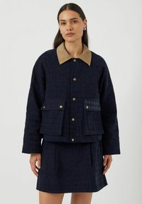Marinblå quiltad jacka med en beige kordflöjelkrage, två framfickor och houndstooth-detalj på en ficka. Slät textur, kortad design.