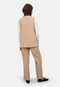 Gilet beige sovrapposto a una camicia a maniche lunghe, abbinato a pantaloni beige coordinati. Il materiale appare morbido, con una texture liscia. Foulard marrone.