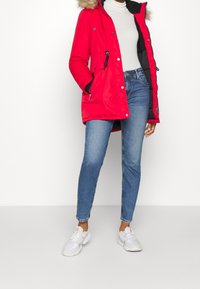 Röd isolerad parkas med huva i päls, dragkedja fram och tryckknappsstängning. Kombinerad med ljusblå jeans och vita sneakers.