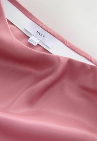 Ružičasta tkanina s glatkom teksturom, koja ima bijeli ovratnik i oznaku s natpisima "NEXT" i "MADE IN MOROCCO." Prikaz iz blizine.
