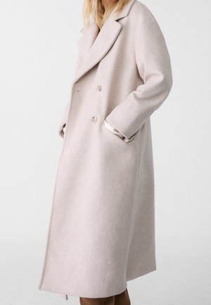 Vrouw die een lange, dubbele rij beige wollen jas draagt met brede revers en handen in de zakken, tegen een effen witte achtergrond.