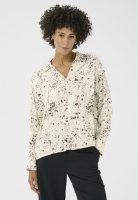 Een vrouw met krullend haar draagt een crèmekleurige blouse met bruine spikkels, gecombineerd met zwarte broeken, terwijl ze tegen een eenvoudige witte achtergrond staat.