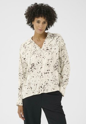 Una donna con capelli ricci indossa una blusa color crema con macchie marroni, abbinata a pantaloni neri, in piedi davanti a uno sfondo bianco uniforme.