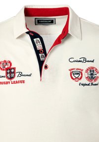 Hvid polo t-shirt med rød krave, marineblå detaljer, broderede logoer og tekst. Har tre knapper ved halsen og er lavet af bomuldsmateriale.