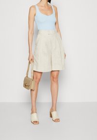 Débardeur bleu clair côtelé, short en lin beige avec plis, mules à talons nude, et un petit sac à main beige avec un accent doré.