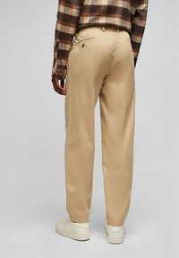 Beige Hose aus leichtem Stoff mit geradem Schnitt, ausgestattet mit Gesäßtaschen und einer glatten Textur. Weiße Sneakers runden den Look ab.