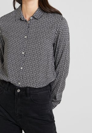 Kvinde iført sort knap-op skjorte med hvidt gentaget tekst "LOVE" og sorte jeans mod en ensfarvet lys baggrund.