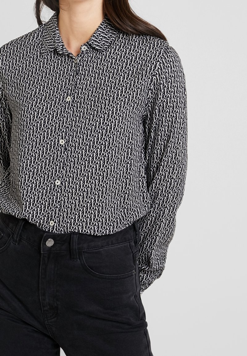 Femme portant une chemise noire à boutons avec impression de texte blanc répété "LOVE" et jean noir sur un fond clair uni.