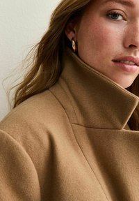 Een tan kleurige wollen jas met een hoge kraag, met een gladde textuur. Het model heeft gouden oorbellen en zacht, golvend haar.