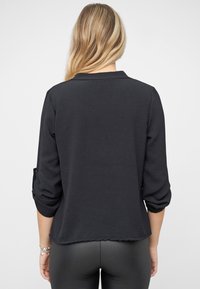 Schwarzes Langarmoberteil mit Kragen, ausgestattet mit einer strukturierten Oberfläche und hochgekrempelten Ärmeln, der Saum ist geschwungen und leicht geschliffen.