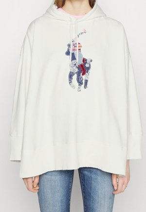 Sweat à capuche blanc oversize avec un motif de joueur de polo multicolore sur la poitrine, porté avec un jean en denim bleu.