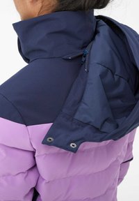 Gepolsterte Jacke mit navy blauem Oberteil und helllila Unterteil, ausgestattet mit einem hohen Kragen und verstecktem Reißverschluss. Glatte Textur und minimale Hardware.