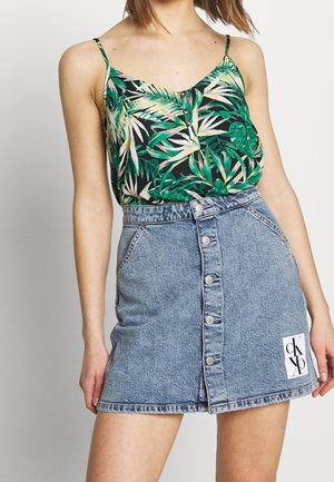 Woman wearing a green and black tropical print sleeveless top and a button-front blue denim mini skirt with a white logo patch.