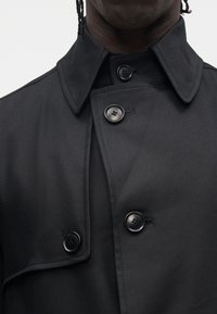 Czarny trencz z ostro zakończonym kołnierzem, dwurzędowym zapięciem i błyszczącymi guzikami. Tkanina wydaje się gładka, o matowym wykończeniu.