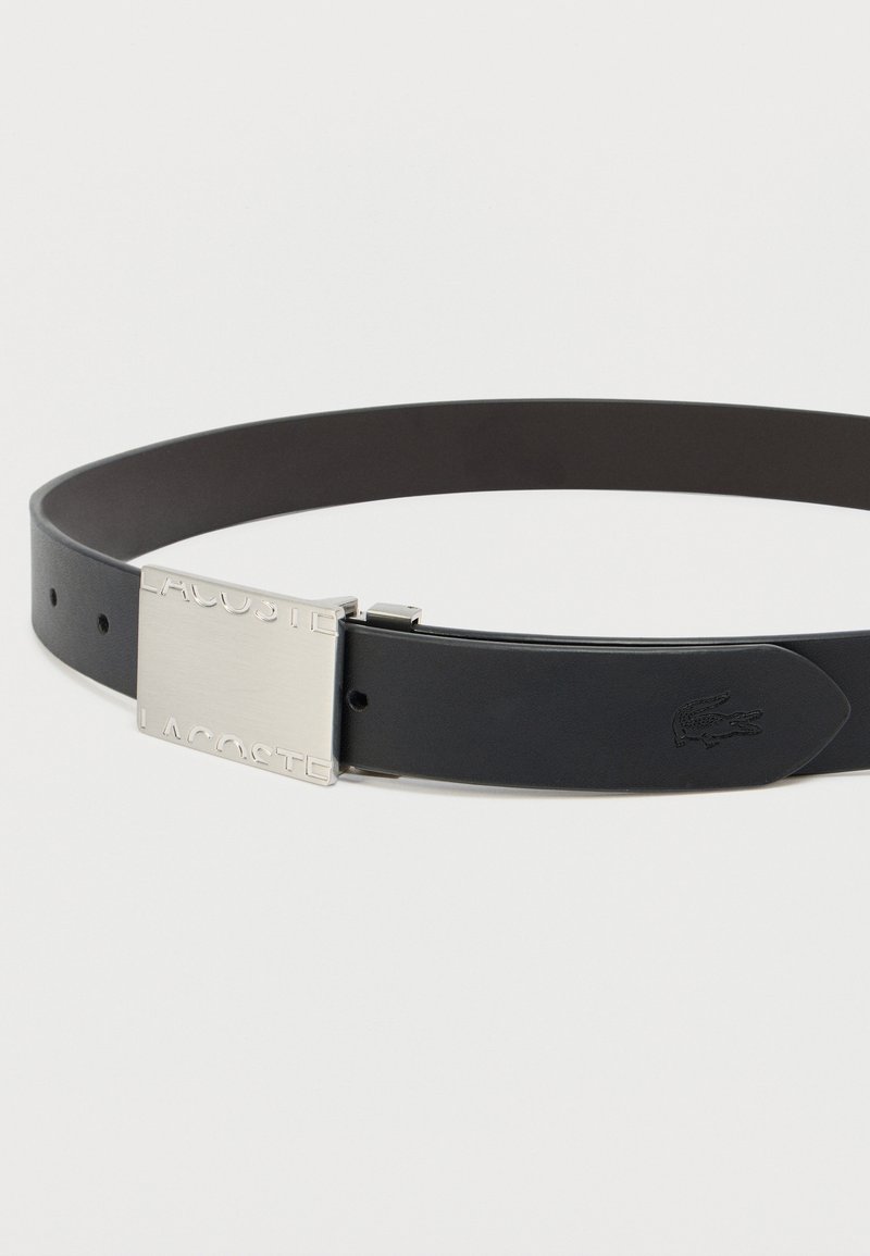 Ceinture en cuir noir avec une boucle rectangulaire en argent portant le logo gravé "LACOSTE" et un emblème de crocodile à l'extrémité.
