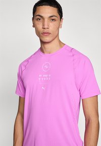 Ροζ αθλητική μπλούζα με κοντά μανίκια και υφή υφάσματος, με λευκό λογότυπο και κείμενο. Διαθέτει μικρές οπές αερισμού στο πλάι.