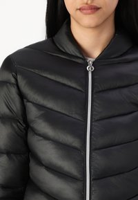 Czarna kurtka puchowa z przeszyciami w kształcie jodełki, wysokim kołnierzem i srebrnym zamkiem błyskawicznym. Tkanina ma gładką, błyszczącą teksturę.