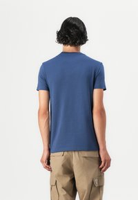 Polo scurt de culoare bleumarin, realizat din bumbac moale, cu guler rotund și textură netedă, prezentat din spate cu pantaloni cargo într-o nuanță bej.
