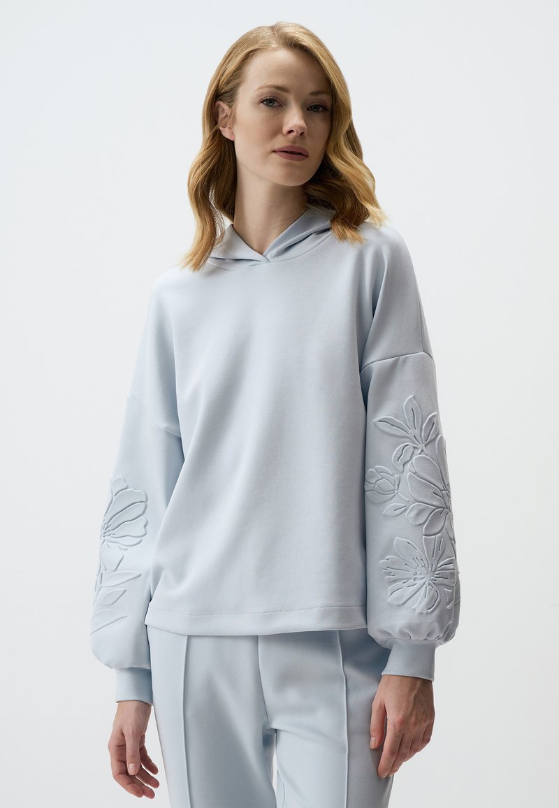 Hoodie bleu clair à coupe décontractée, avec des manches oversized et des motifs floraux en relief sur les manches. Texture douce et design décontracté.