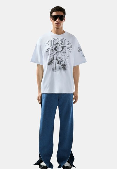 Homme portant un t-shirt blanc oversize avec un graphique de squelette et le texte "GCDS", un jean large bleu avec des ourlets fendus, des lunettes de soleil noires et des chaussures noires.