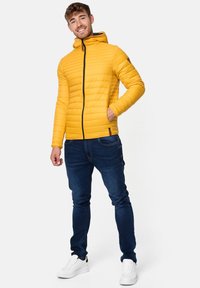 Geel gewatteerde jas met een ritssluiting aan de voorkant en capuchon, met diagonale quiltstructuur en zwarte accenten. Draagt met donkere spijkerbroek en witte sneakers.