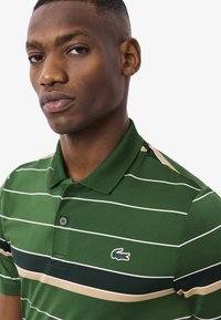 Polo shirt a righe verdi con strisce orizzontali bianche e beige, caratterizzato da un logo di coccodrillo verde sul petto e un design di colletto classico.