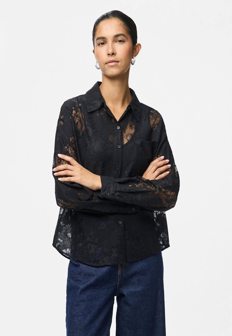 Woman with arms crossed wearing a black lace button-up shirt and blue jeans against a light gray background.