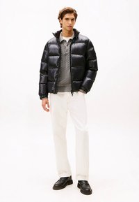 Black puffer jacket with horizontal quilting, high collar, front zipper, worn over a gray cable-knit sweater and white pants. Black loafers complete the look.