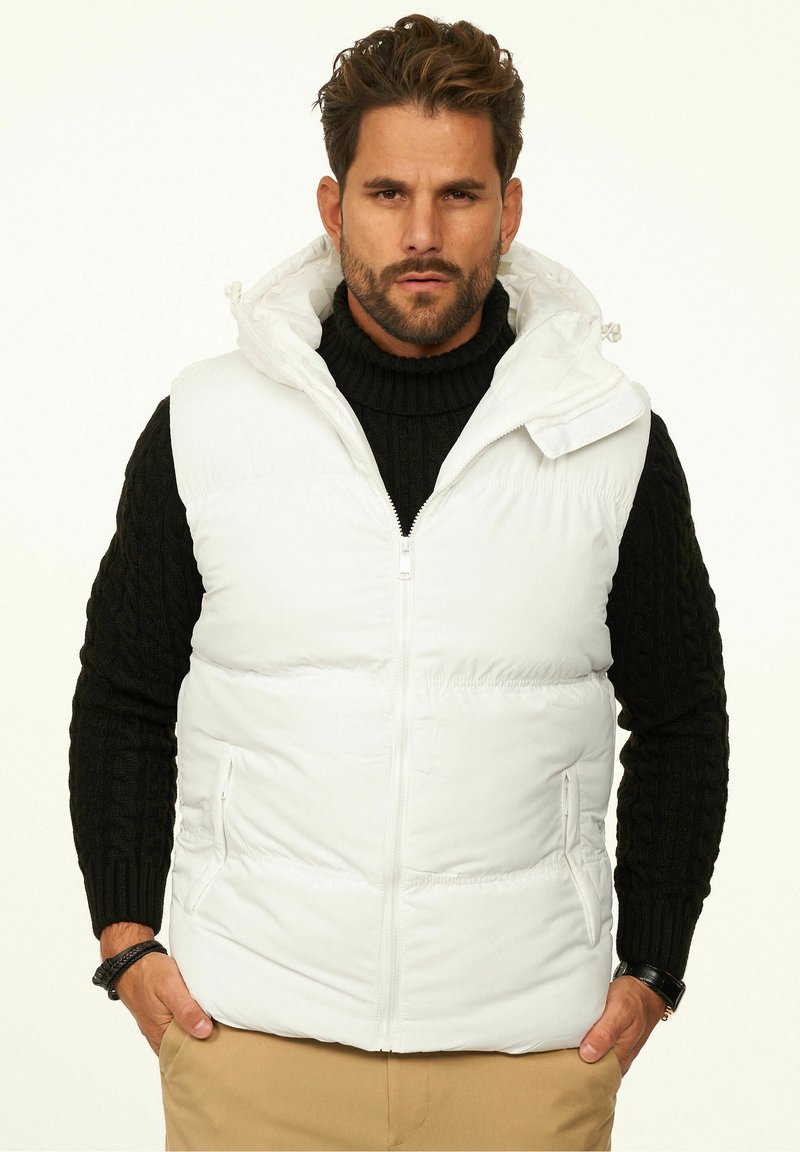 Weiße wattierte Weste mit Kapuze, Reißverschluss vorne, Seitentaschen und einem strukturierten schwarzen Pullover darunter. Das Modell steht vor einem hellen Hintergrund.