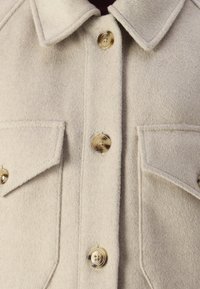 Beige villasegu segu robe, millel krae, kaks rinna taskut ja suured, ümmargused dekoratiivnuppud, millel on sile pind.