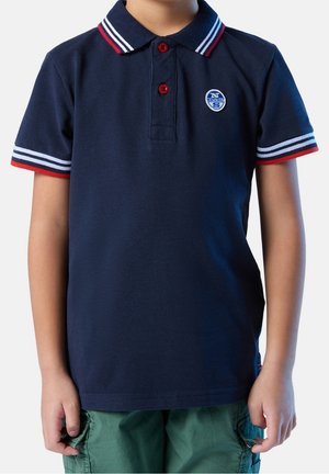 Lapsi pukeutuneena tummansiniseen poolopaitaan, jossa on punavalkoraidalliset kaulus ja hihat, yhdistettynä vihreisiin cargoshortseihin.